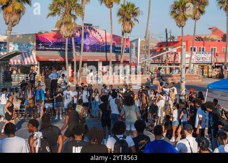 Eine vielfältige Menschenmenge versammelt sich um einen Basketballplatz in Venice Beach, Los Angeles. Farbenfrohe Wandgemälde, Palmen und eine Werbetafel mit Riesenrad runden das ab Stockfoto