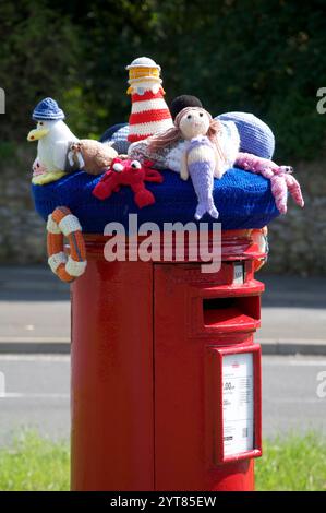 Ein farbenfroher Strickmütze aus Wolle mit Meeresmotiv und gehäkelten Möwen, Meerjungfrauen und anderen Meeresbewohnern ziert eine traditionelle rote Säulenkasten. UK. Stockfoto