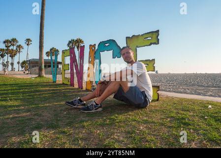 Ein Mann in lässiger Kleidung sitzt auf Gras neben einem lebendigen Venedig-Schild. Der Hintergrund ist ein Sandstrand, ein klarer blauer Himmel und verstreute Palmen. Stockfoto