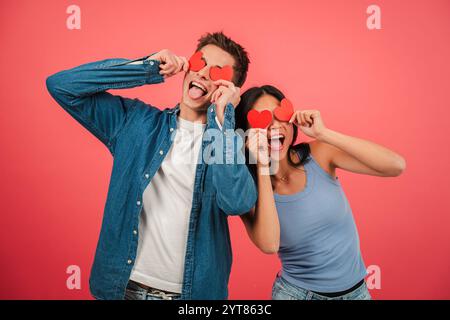 Verspieltes junges Paar mit roten Herzformen und lustigen Gesichtern vor leuchtendem rotem Hintergrund, das Liebe und Glück mit fröhlichen Ausdrücken in einem lustigen und kreativen Valentinstag feiert. Hochwertige Fotos Stockfoto