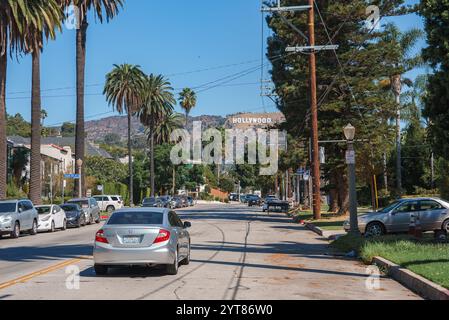 Eine Straße in Los Angeles zeigt das Hollywood-Schild in den Hügeln, gesäumt von hohen Palmen. Die Autos parken an den Seiten, und ein silbernes Auto fährt. Stockfoto