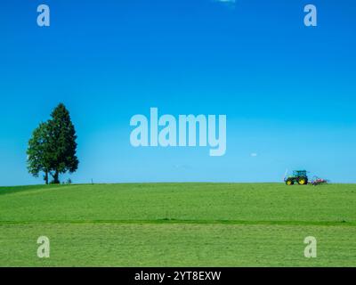 Landschaftsansicht einer frisch gemähten Weide mit Einzelbaum und Traktor mit Mähanhänger unter blauem Himmel in Bayern. Stockfoto