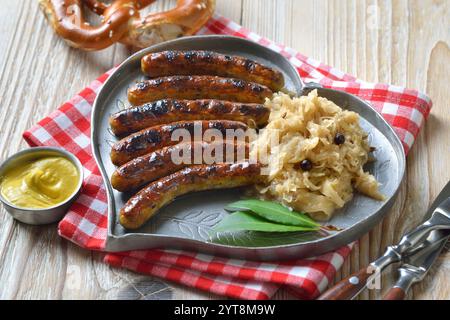 Sechs gegrillte Nürnberger Würstchen mit Sauerkraut auf einer herzförmigen Zinnplatte, serviert mit Senf und Brezel Stockfoto