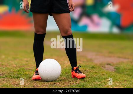 Ansicht der Beine einer brünetten Fußballspielerin, die zum Kicken des Fußballs läuft. Auf einem grünen Gras draußen, mit farbenfrohem Hintergrund. Mit Sportbekleidung. Stockfoto