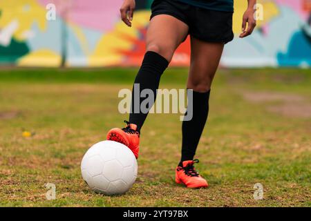 Ansicht der Beine einer brünetten Fußballspielerin, die zum Kicken des Fußballs läuft. Auf einem grünen Gras draußen, mit farbenfrohem Hintergrund. Mit Sportbekleidung. Stockfoto