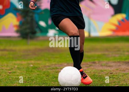 Ansicht der Beine einer brünetten Fußballspielerin, die zum Kicken des Fußballs läuft. Auf einem grünen Gras draußen, mit farbenfrohem Hintergrund. Mit Sportbekleidung. Stockfoto