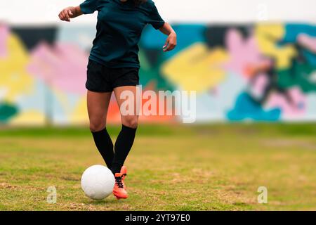 Ansicht der Beine einer brünetten Fußballspielerin, die zum Kicken des Fußballs läuft. Auf einem grünen Gras draußen, mit farbenfrohem Hintergrund. Mit Sportbekleidung. Stockfoto