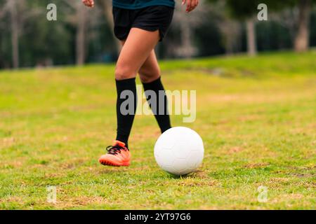 Ansicht der Beine einer brünetten Fußballspielerin, die zum Kicken des Fußballs läuft. Auf einem grünen Gras draußen, mit farbenfrohem Hintergrund. Mit Sportbekleidung. Stockfoto