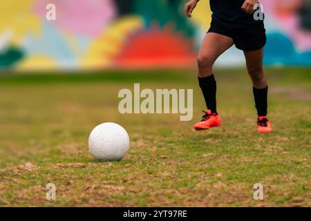 Ansicht der Beine einer brünetten Fußballspielerin, die zum Kicken des Fußballs läuft. Auf einem grünen Gras draußen, mit farbenfrohem Hintergrund. Mit Sportbekleidung. Stockfoto