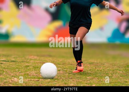 Ansicht der Beine einer brünetten Fußballspielerin, die zum Kicken des Fußballs läuft. Auf einem grünen Gras draußen, mit farbenfrohem Hintergrund. Mit Sportbekleidung. Stockfoto