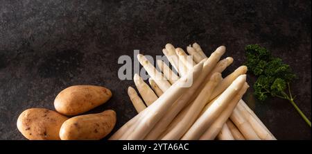 Frischer weißer Spargel und rohe Kartoffeln auf einem modernen schwarzen Flagstein. Saisonales Gemüse für die Zubereitung eines gesunden Spargelmenüs. Sonnige Küchen-Szene für die Frühjahrs-Gastronomie. Stockfoto
