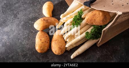 Frischer weißer Spargel und rohe Kartoffeln in einer Einkaufstasche. Saisonales Frühlingsgemüse auf modernem schwarzem Schiefer. Sonnige Küchenszene für ein gesundes Gastronomie-Konzept mit Platz für Text. Stockfoto