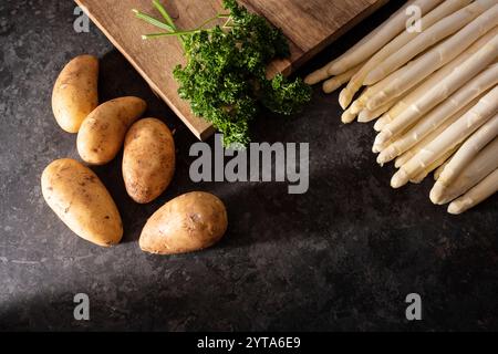 Frischer weißer Spargel und rohe Kartoffeln mit Holzschneidebrett saisonales Frühlingsgemüse auf modernem schwarzen Schiefer. Sonnige Küchenszene für ein gesundes Gastronomie-Konzept mit Platz für Text. Stockfoto