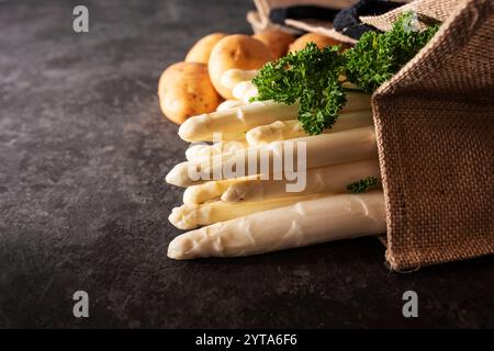 Frischer weißer Spargel und rohe Kartoffeln in einer Einkaufstasche. Saisonales Frühlingsgemüse auf modernem schwarzem Schiefer. Sonnige Küchenszene für ein gesundes Gastronomie-Konzept mit Platz für Text. Stockfoto