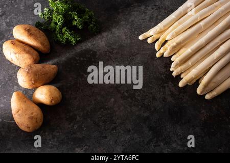Frischer weißer Spargel und rohe Kartoffeln auf schwarzem Hintergrund. Saisonales Frühlingsgemüse auf modernem schwarzem Schiefer. Sonnige Küchenszene für ein gesundes Gastronomie-Konzept mit Platz für Text. Stockfoto