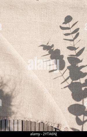 Leinentischdecke im Sonnenlicht mit Blattschatten. Beigefarbene Leinenstoff auf rustikalem Holztisch. Vertikaler Hintergrund für Hochzeits- und Geburtstagsfeiern im Boho-Stil mit Platz für Text. Stockfoto