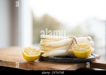 Gebundener weißer Spargel mit Zitronenscheiben auf Holzschneidebrett und schwarzer Keramikplatte, vor einem Küchenfenster mit geringer Schärfentiefe. Küche für saisonale Gastronomie. Stockfoto