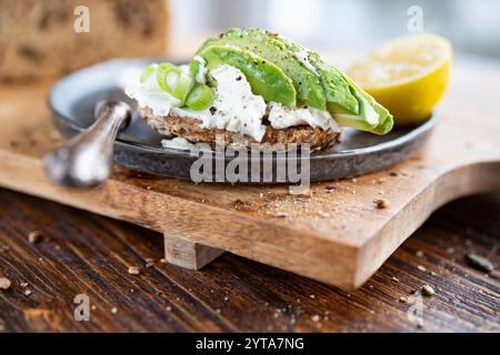 Frisches Mehrkornbrot mit Frischkäse und Avocado auf schwarzer Keramikplatte. Nahaufnahme vor einem hellen Fenster. Kurze Schärfentiefe. Stockfoto