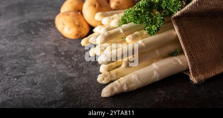 Frischer weißer Spargel und rohe Kartoffeln in einer Einkaufstasche. Saisonales Frühlingsgemüse auf schwarzem Schiefer. Saisonal und gesundes Gastronomie-Konzept mit Platz für Text. Stockfoto