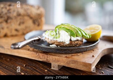 Frische Avocadoscheiben auf Getreidebrot mit Frischkäse und Zitrone. Vegetarische Speisen auf hölzernem Schneidebrett. Nahaufnahme vor einem hellen Fenster. Kurze Schärfentiefe. Stockfoto