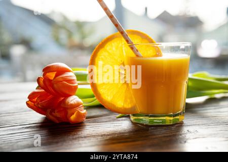 Glas frisch gepresster Orangensaft mit einer Scheibe Orange und einem Strauß Tulpen auf einem Holztisch. Nahaufnahme. Stockfoto