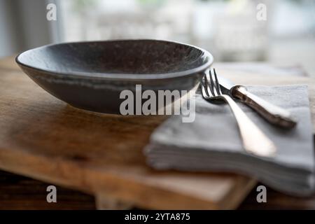 Einfache rustikale Tischeinstellung auf hölzernem Schneidebrett. Altes silbernes Besteck mit Leinenserviette und schwarzem Keramikgeschirr. Stockfoto