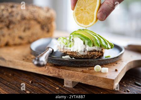 Frische Avocadoscheiben auf Getreidebrot mit Frischkäse. Hand von oben mit Zitrone. Vegetarische Speisen auf hölzernem Schneidebrett. Nahaufnahme vor einem hellen Fenster. Stockfoto