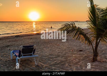 Eine Liege sitzt auf dem Sand neben einer Palme. Die Sonne untergeht im Hintergrund und schafft eine warme und entspannende Atmosphäre Stockfoto