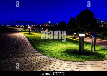 Ein Park mit einer Brücke im Hintergrund und einem Schild auf der rechten Seite. Die Lichter auf dem Gras und die Straßenlaternen schaffen eine friedliche und ruhige Atmosphäre Stockfoto