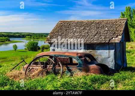 Ein verrostetes altes Auto parkt vor einem kleinen blauen Haus. Das Auto ist von Gras umgeben und das Haus liegt auf einem Hügel mit Blick auf einen Fluss Stockfoto