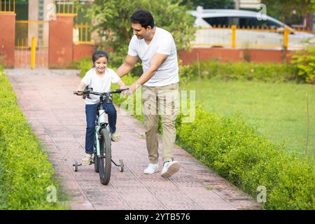 Der junge indische Vater bringt ihrer Tochter bei, im Park im Freien Fahrrad zu fahren. Dad hilft und unterstützt sie beim Radfahren und Treten. Stockfoto