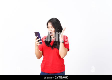Schockierendes Wow-Gesicht, das Smartphone ansieht und Smartphone der schönen asiatischen Frau hält, die ein rotes Hemd trägt, isoliert auf weißem Hintergrund Stockfoto