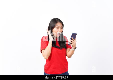 Schockierendes Wow-Gesicht, das Smartphone ansieht und Smartphone der schönen asiatischen Frau hält, die ein rotes Hemd trägt, isoliert auf weißem Hintergrund Stockfoto