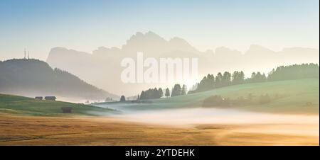 Dieses Foto zeigt die Seiser Alm in den Dolomiten bei Sonnenaufgang mit Nebel über grüne Wiesen, verstreute Hütten und weit entfernte Berge Stockfoto