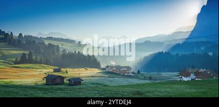 Dieses Foto zeigt die Seiser Alm in den Dolomiten bei Sonnenaufgang mit Nebel über grüne Wiesen, verstreute Hütten und weit entfernte Berge Stockfoto