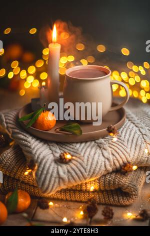 Eine Tasse heißes Getränk steht auf einem Tablett mit Mandarine-Orange und brennenden Kerzen, auf einer gemütlichen Strickdecke mit warmer, einladender Beleuchtung, cre Stockfoto