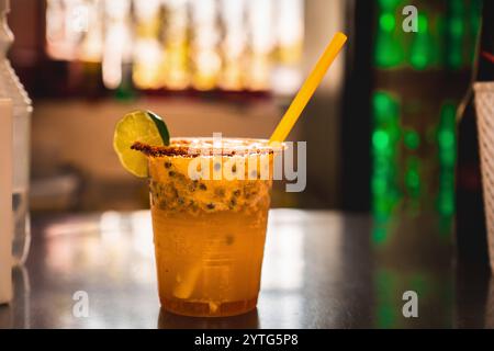 Eine tropische kolumbianische Michelada mit Bier, frischer Passionsfrucht, einem mit Chili gesalzenen Rand und einer Limettenscheibe. Serviert in einem Plastikbecher mit lebendigem Hintergrund, Stockfoto