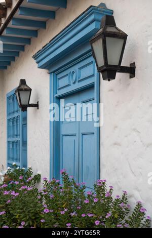 Wunderschöne blaue hölzerne Tür im Kolonialstil mit dekorativen Laternen, weißen Wänden und lila Blumen im Vordergrund, die eine charmante Tradition einfangen Stockfoto