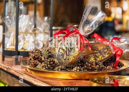 Nahaufnahme von eingewickelten dekorativen Lebkuchenkeksen mit roten Bändern, angeordnet auf einem goldenen Tablett mit Tannenzapfen. Weihnachtsmarkt, Riga, Lettland Stockfoto