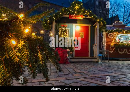Festliche Santa Claus Hütte mit einer knallroten Tür, Girlandendekorationen und Weihnachtsbaum umgeben von Geschenken, beleuchtet von warmen Feenlichtern Stockfoto