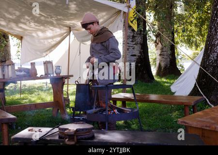 Mantova, Italien - 15. September 2024: Junge in mittelalterlicher Kleidung neben einem Feuerherd in einem Lager, der ein Getränk umrührt, das kocht Stockfoto