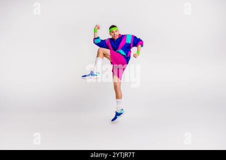 Energiegeladener junger Mann in farbenfroher Retro-Sportbekleidung, der fröhlich in weißem Studio-Hintergrund springt Stockfoto