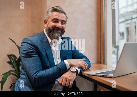Ein glücklicher, reifer Geschäftsmann, der im Büro oder Café am Laptop arbeitet, eine Kamera ansieht und lächelt. Stockfoto