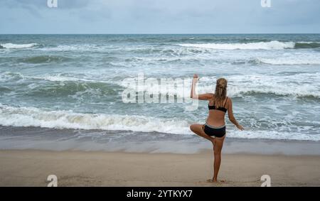 Eine Frau übt Yoga an einem Sandstrand in Thailand, mit Blick auf den Ozean. Wellen krachen unter bewölktem Himmel gegen die Küste und schaffen eine friedliche und dennoch friedliche Dynam Stockfoto