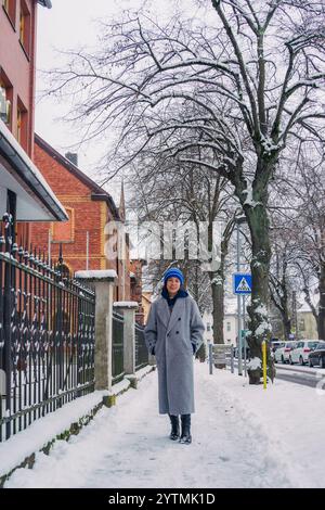 Eine Frau steht auf einer verschneiten Straße, trägt einen grauen Mantel und einen bunten Hut und hält einen ruhigen Wintermoment fest. Stockfoto