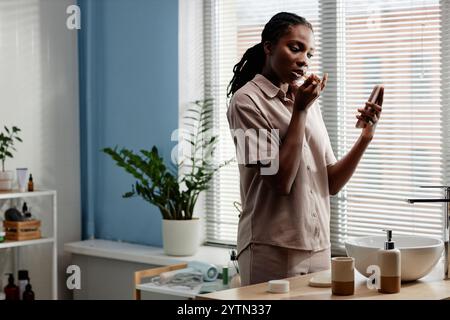 Mittelgroße Aufnahme einer jungen schwarzen Frau, die Zähne putzt und während der morgendlichen Selbstpflegeroutine im Badezimmer Textnachrichten am Telefon übermittelt, Kopierraum Stockfoto