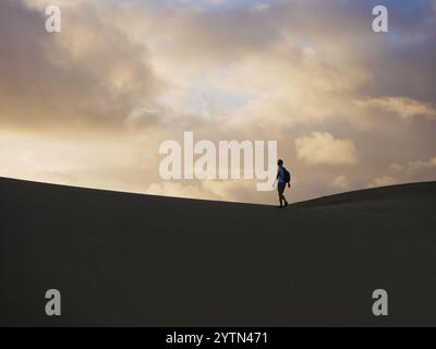 Ein Reisender Tourist mit Rucksack spaziert bei Sonnenuntergang durch die Wüste. Dünen in Maspalomas, Gran Canaria, Spanien. Stockfoto