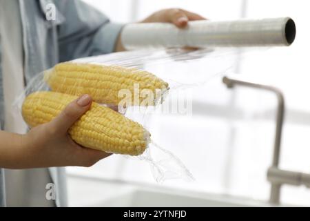 Frau, die Plastikfutterverpackung über Maiskolben in der Küche legt, Nahaufnahme Stockfoto