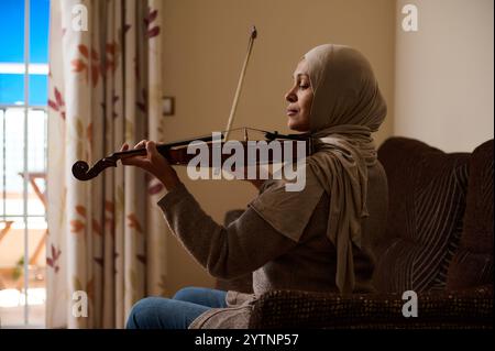 Eine Frau im Hijab spielt Geige, sitzt auf einer Couch in einem warm beleuchteten Wohnzimmer, umgeben von Blumenvorhängen. Die Szene strahlt Frieden aus, Harmonie, und Stockfoto