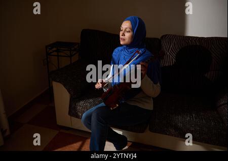 Eine Frau mit blauem Hijab sitzt auf einer Couch und spielt geschickt ihre Violine, wobei Licht und Schatten ihren fokussierten Ausdruck hervorheben. Stockfoto